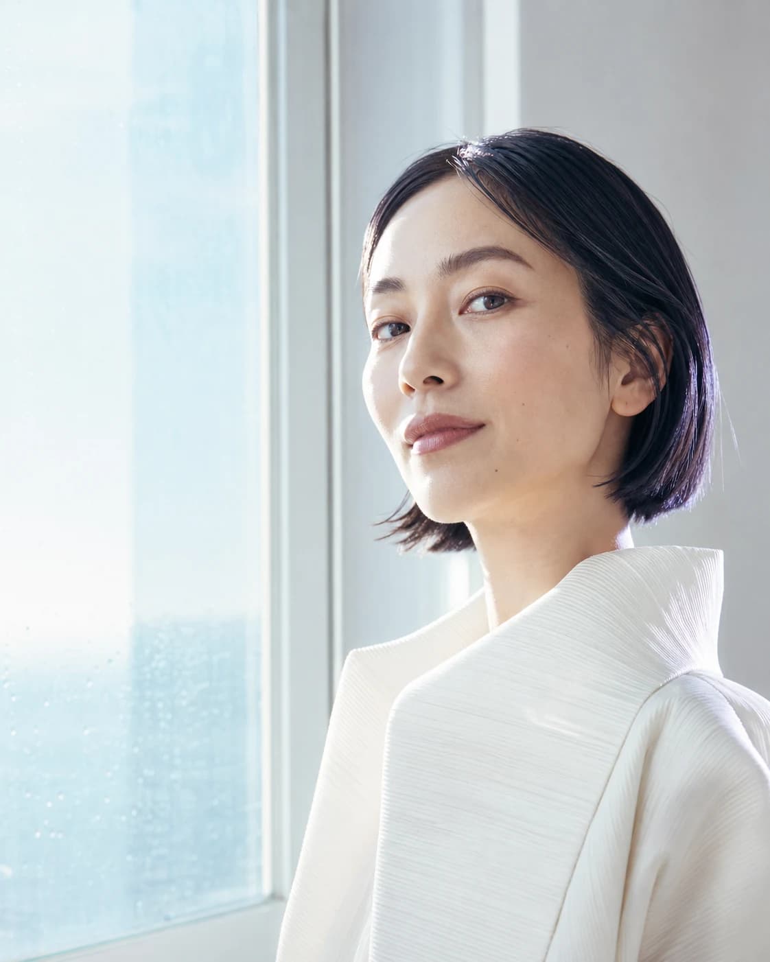 Bright editorial portrait of an East Asian woman in an ivory jacket beside a sunlit window